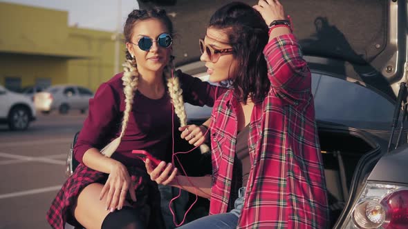 Two Young Attractive Girlfriends in Sunglasses Sitting Inside of the Open Car Trunk in the Parking