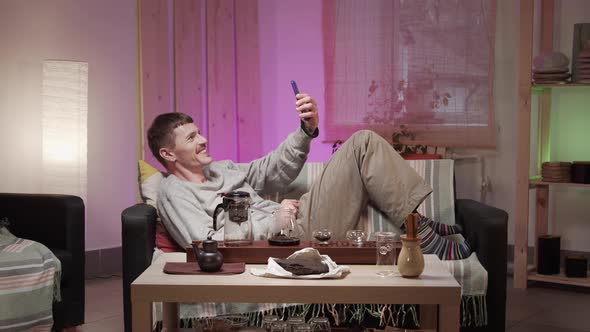 A Young Man with a Mustache Is Lying on a Sofa in a Room on Self-isolation and Communicating with