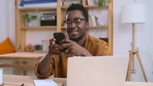African Man Holding Phone in Hand Next to Laptop in Home White Interior Spbas