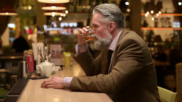 Gray-haired adult man with a beard sits alone in a bar drinking whiskey on ice