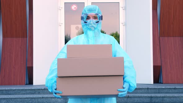 Courier, in Protective Uniform, Mask, Goggles, Holds in Hands Some Cardboard Boxes, on Background of