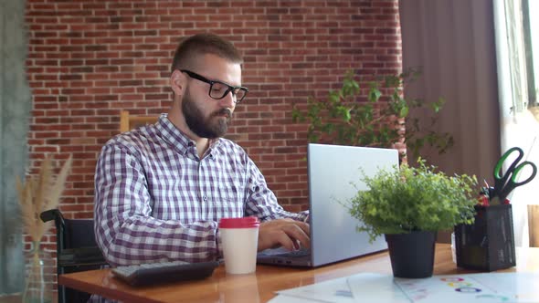 Disabled Person Man with Glasses is Sitting in Wheelchair and Typing on Laptop Turns His Head and