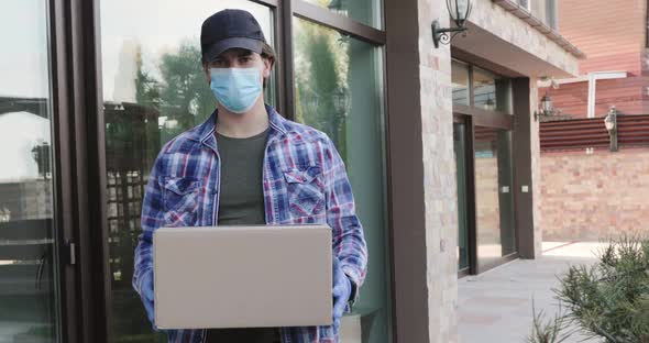 Man Wore in Mask and Gloves with Parcel in Hands at House Looks at Camera