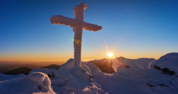 A frozen Christian cross in the winter mountains by the setting sun. Sunset