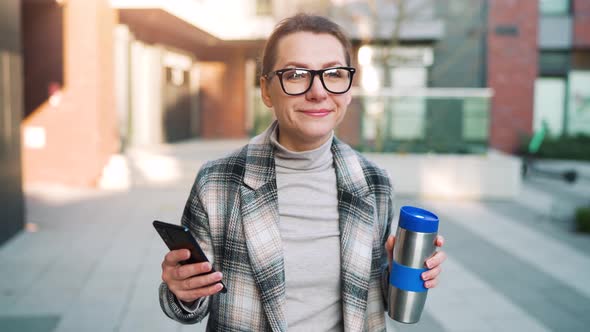 Caucasian Businesswoman with Glasses and a Coat Walks Through the Business District with Thermo Cup