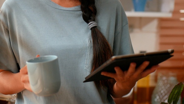 Young Asian woman using tablet beside a window in kitchen at home and drinking coffee.