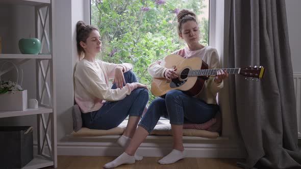 Wide Shot Talented Beautiful Twin Sisters Sitting on Windowsill Playing Guitar Singing