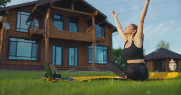 Young Woman in Sports Clothes Does Yoga Exercises Using Laptop in the Backyard on the Green Grass