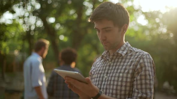 Young Handsome Caucasian Male in Plaid Shirt Standing in Park with Tablet Showing Ok with Thumb with