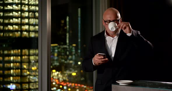 a Middle-aged Man in Glasses and a Suit Checks His Phone and Drinks Coffee By the Big Window