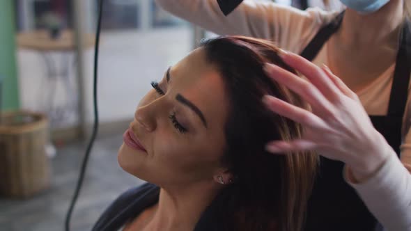 Female hairdresser wearing face mask blow drying hair of female customer at hair salon