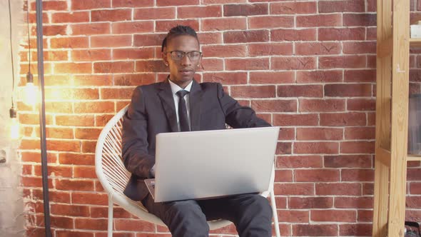 Serious black man in an official suit and glasses uses laptop, sitting on a chair against