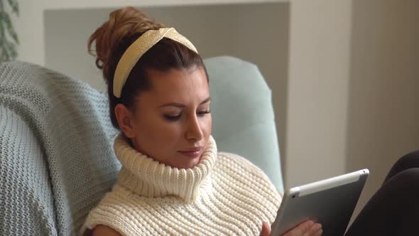 beautiful young woman in pajamas at home in a soft armchair works on the digital tablet