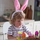 Cute two-year-old boy in bunny ears paint Easter eggs with multi-color paints sitting at the table - VideoHive Item for Sale