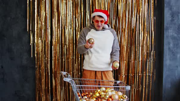 Young Male with Shopping Cart Filled of Assorted Baubles Standing Against Wall with Golden Tinsel