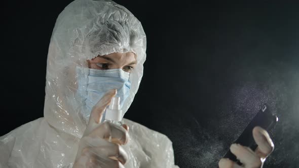 Man in a Medical Mask and Protective Suit Is Spraying an Antiseptic with a Mobile Phone on a Dark