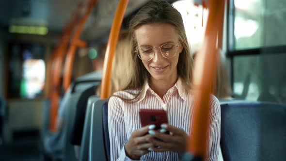 A Smiling Young Woman is Using Smartphone Social Media Reading Messages in a Bus and Smiling