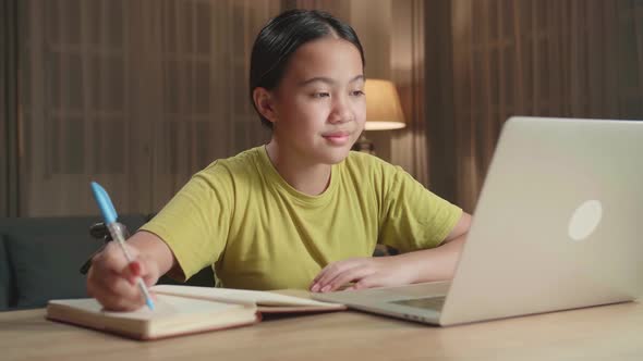 Asian Kid Girl Sitting In A Wheelchair While Using Laptop Computer And Note In Book At Home