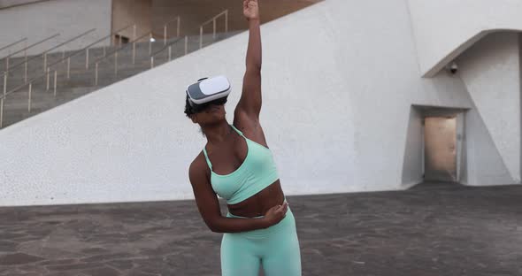Fit african woman doing sport stretching exercise with virtual reality goggles in the city