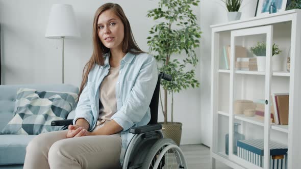 Portrait of Young Disabled Woman Sitting in Wheelchair at Home and Looking at Camera with Light
