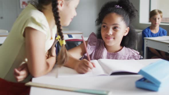 Video of happy diverse girls at desk doing lessons together in classroom