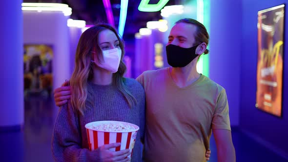 Happy Young Caucasian Couple in Mask Go to the Cinema Walking By Hall with Bucket of Popcorn