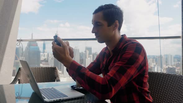 Busy Business Man Uses Smartphone Works at a Laptop and Drinks Coffee in a Cafe on the High Floor of