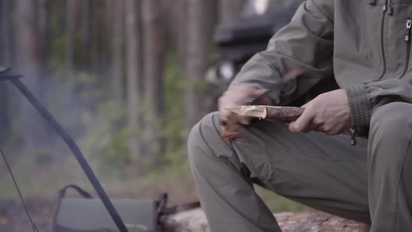 Male Tourist Is Sitting on Log and Planing Wooden Peg with Knife in Summer Forest. Cooking Dinner on