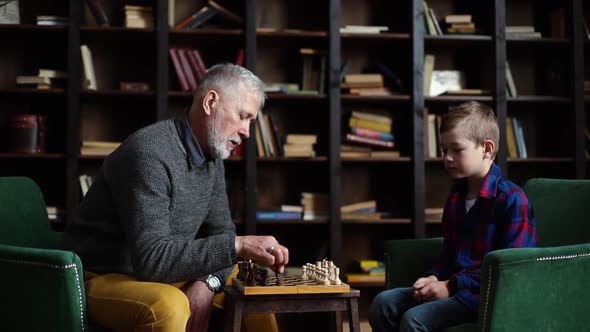 Loving Old Senior Grandfather and Grandson Are Playing Chess Sitting at Desk in Cozy Living Room