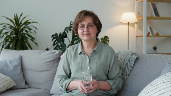 Beautiful Old Lady Looking in Camera Sitting in the Living Room of Her House with a Glass of Water