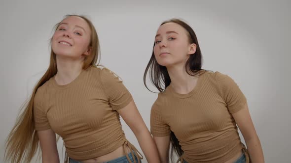 Slim Beautiful Identical Twin Sisters Dancing Simultaneously at Grey Background Looking at Camera