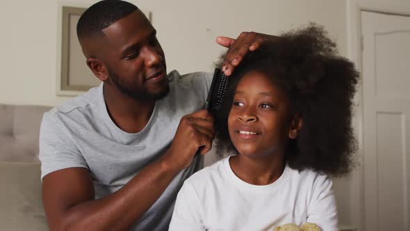 African american father brushing his daughters hair while sitting on the bed at home