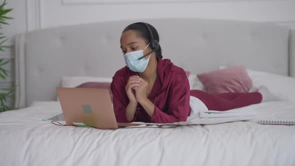 African American Young Woman in Coronavirus Face Mask Lying on Bed Discussing Business Idea Online