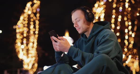 Young Man Using Phone in Headphones in Evening City
