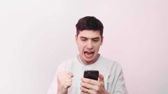 A Pleased Young Man is Using His Smartphone While Doing Winner Gesture Standing Ion Pink Background