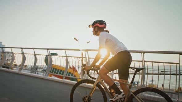 Female in Sportswear and Protective Helmet is Riding a Sports Bike Uphill Along Bridge Near a Sea