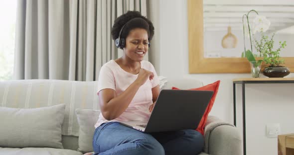 Happy african american woman wearing headphones sitting on sofa, using laptop