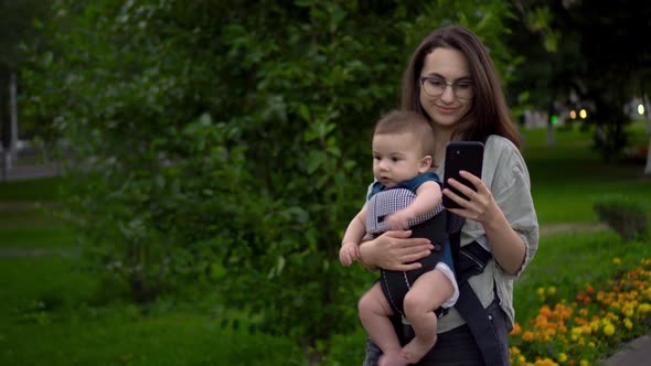 A Young Woman Walks Through the Park with a Child and a Phone in Her Hands