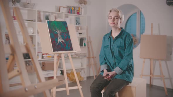 A Young Woman Artist with Short Blonde Hair Sitting in the Art Studio After Finishing the Painting