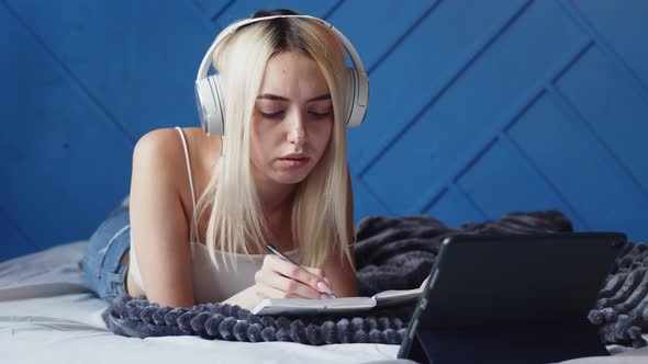 Young Caucasian Woman Student is Lying on Her Bed with Headphones on and Taking Notes While Watching