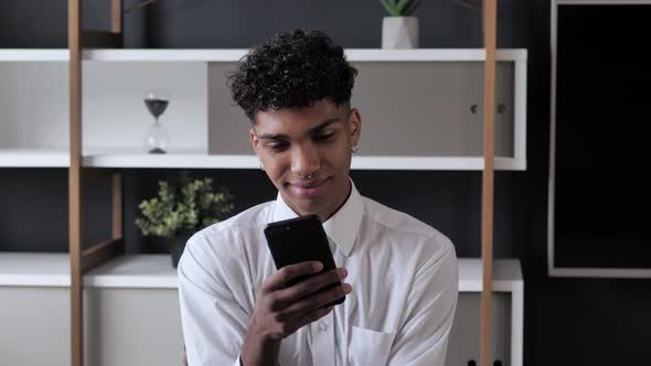 Young African American Student Holding Smartphone Device Texting Sms Message Sitting at Modern