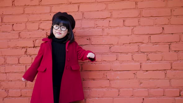 Korean Girl in a Red Coat and Cap and Round Glasses Dance on Red Brick Wall the Street in Autumn