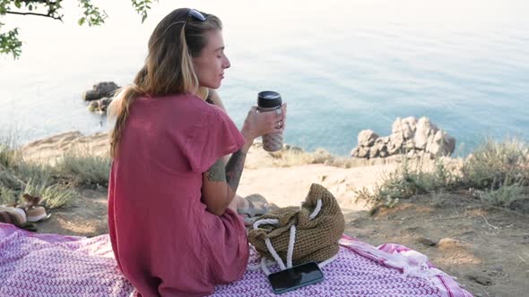 Young Woman Sitting on a Blanket on the Beach By the Sea Looking in Front of Her and Holding a