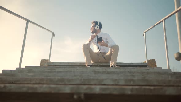 Man Listening To Music In Headphone. Businessman Joyful Sitting On Stairs And Listening Music.