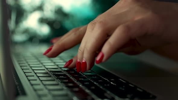 Freelancer Woman with Red Manicure Works on a Laptop Sitting at a Table on a Blurred Background