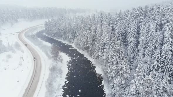 Aerial View of a Country Road in Winter Near a River and Coniferous Forest During Snowfall. The