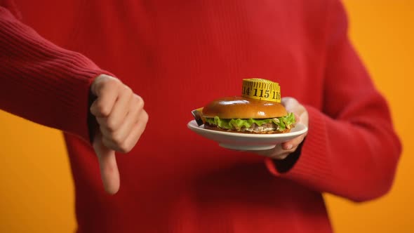 Female Holding Plate With Burger and Measuring Tape Showing Thumbs Down, Diet