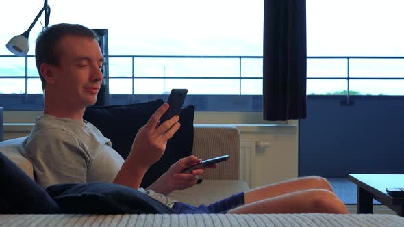 A Young Man Watches Television in Living Room in the Apartment and Works on the Smartphone