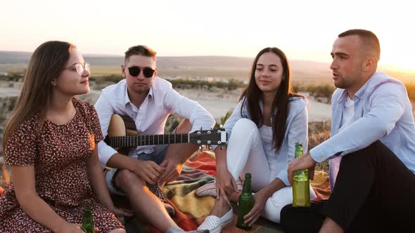 Four Friends Cheering with Beer and Smiling While Sitting on the Blanket at a Summer Picnic