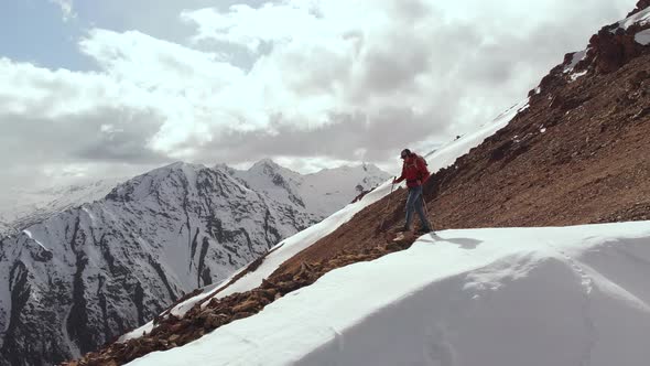 Aerial View of a Young Man in a Cap Sunglasses with a Backpack and Trekking Poles Walking Down the